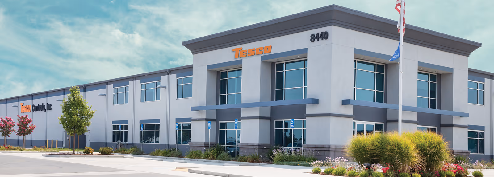 Modern corporate office building with large windows, labeled Tesco and Tecnu Controls, Inc., with landscaping and clear sky.