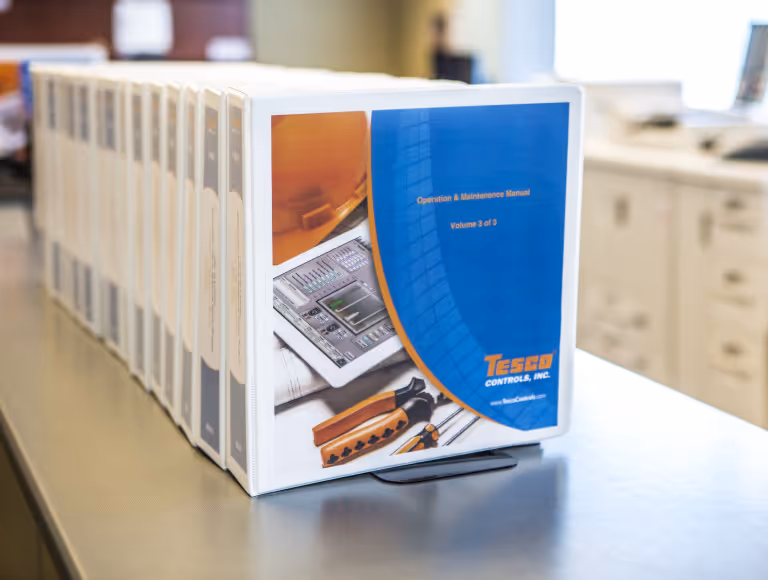 Row of binders on a table with the front one labeled 'Operation & Maintenance Manual Volume 3 of 3' from Tesco Controls, Inc.