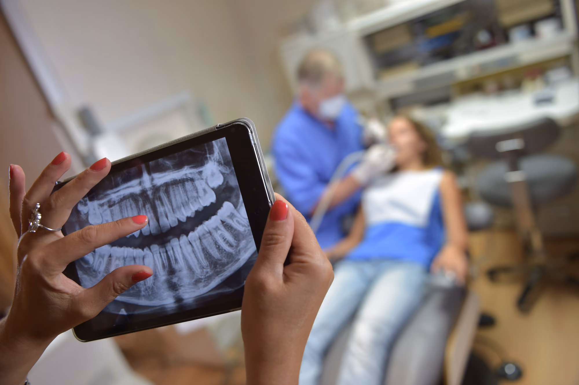 Dental X-ray being reviewed on a tablet in the foreground with patient in treatment chair blurred in background.