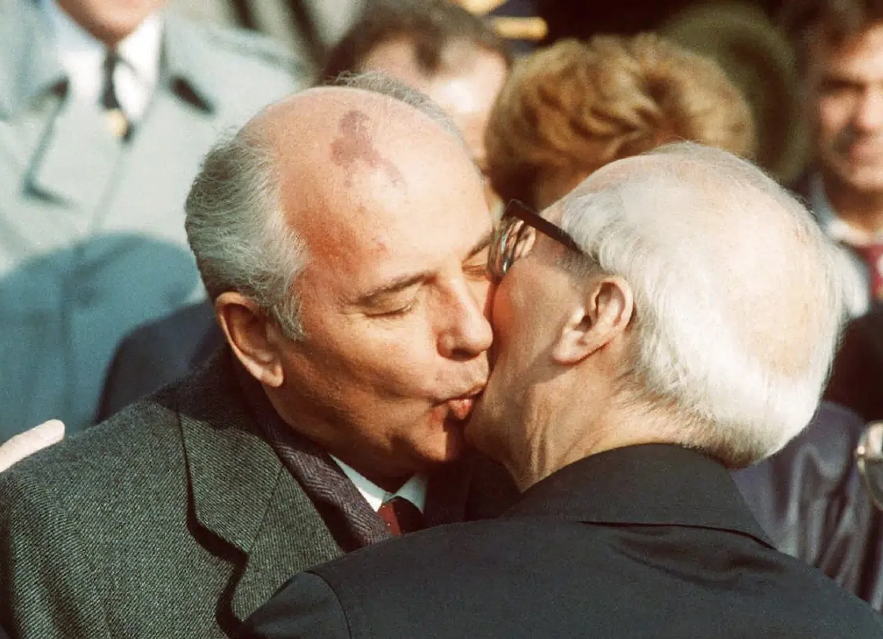 Two older men warmly greeting each other with a kiss on the cheek in a public setting.