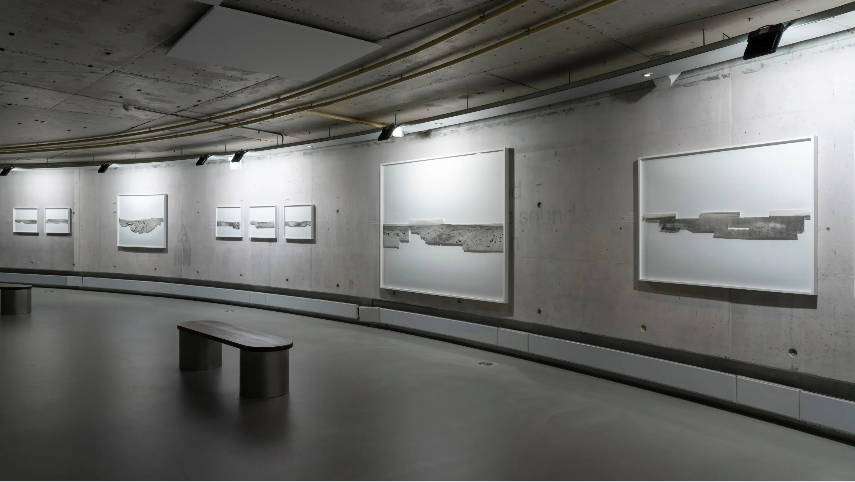 Curved gallery wall with framed abstract artworks featuring horizontal textures, modern concrete ceiling and floor, and two benches.