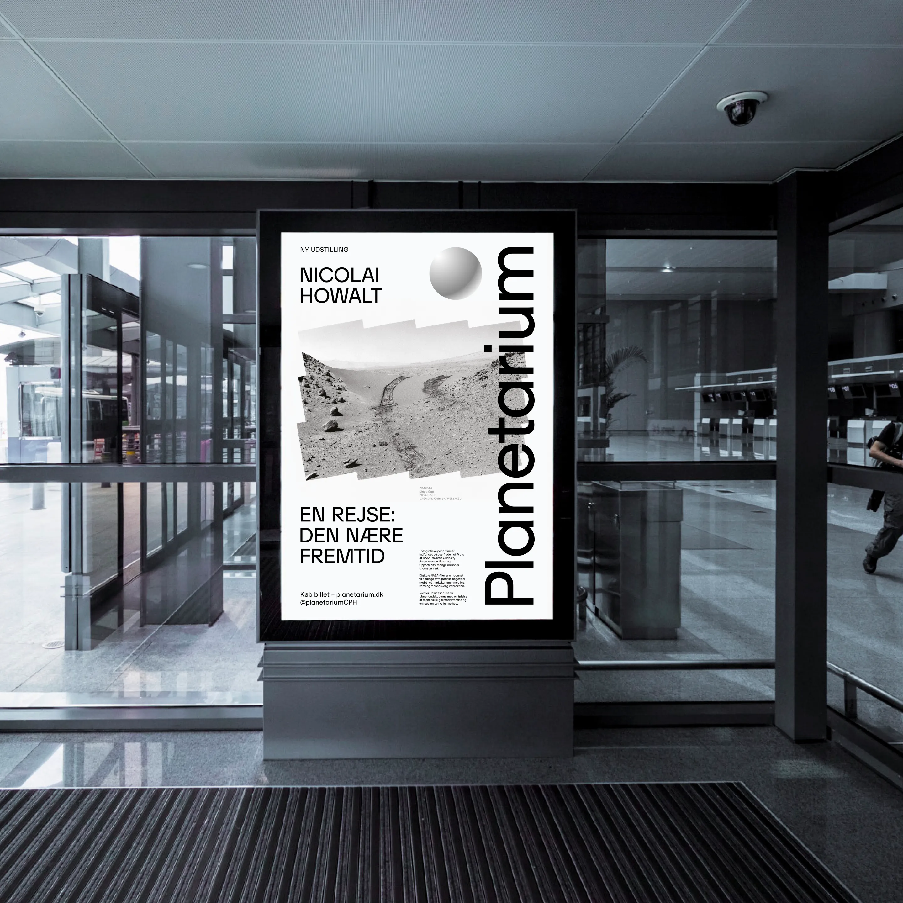 Lit poster in a modern building advertising a new exhibition by Nicolai Howalt at the Planetarium, titled 'En rejse: Den nære fremtid' with a black-and-white landscape photo of rocky terrain.