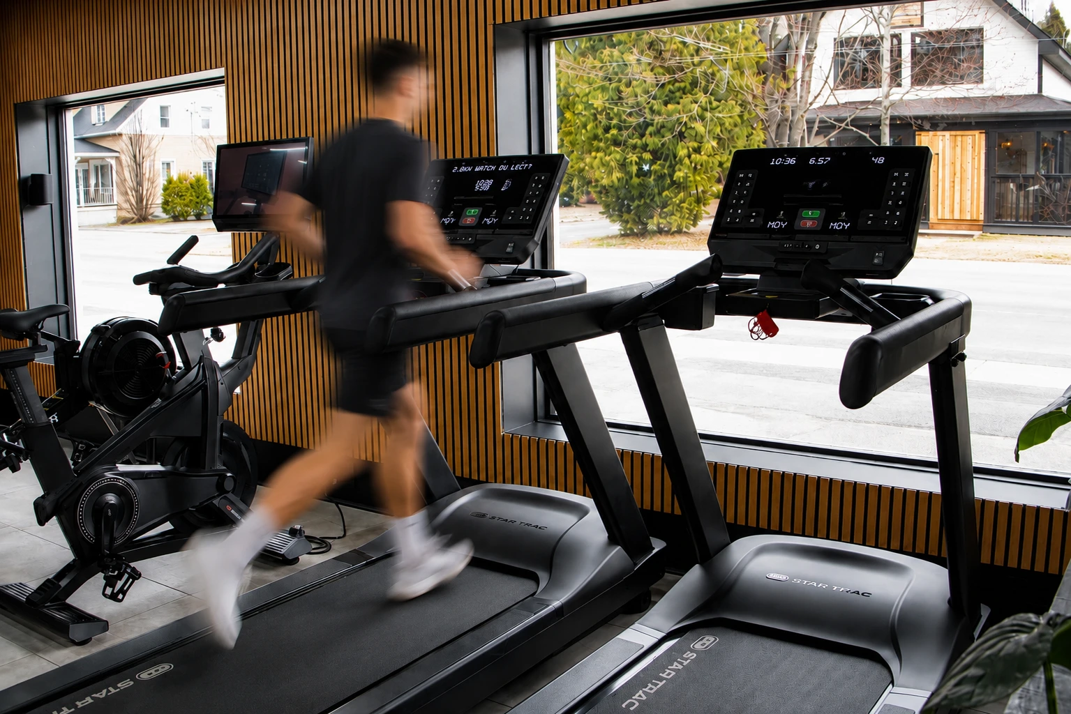 Salle de gym moderne avec appareils de musculation, éclairage LED au plafond et plantes vertes à l'entrée.