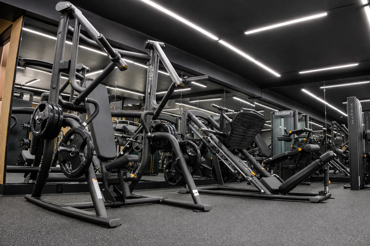 Intérieur d'une salle de sport moderne avec des équipements de musculation, un sol noir avec des éclats, un plafond noir avec des lumières LED blanches en forme de lignes géométriques, et des personnes s'entraînant.