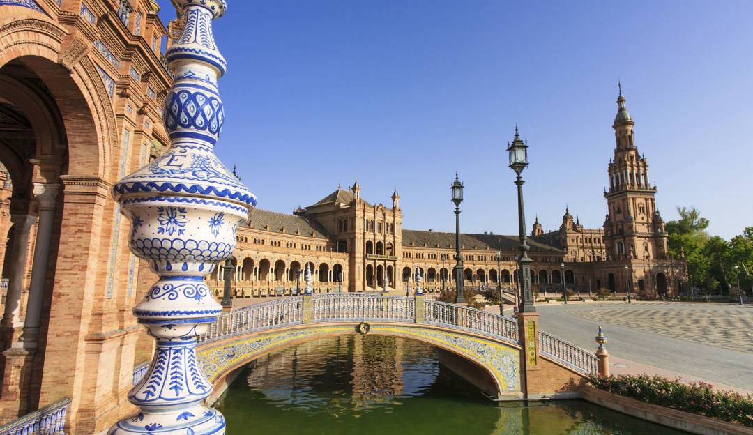 View of a decorative ceramic baluster and a tiled bridge over a canal at Plaza de España in Seville, Spain, with historic buildings and a tower in the background under a clear blue sky.