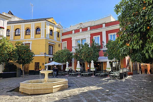 Sunlit cobblestone plaza with yellow and red buildings, outdoor café tables, orange trees, and a stone fountain in the foreground.