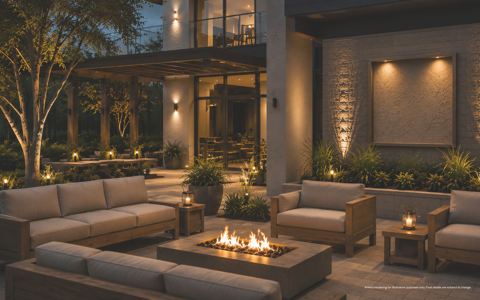 Modern outdoor patio area at dusk with beige cushioned wooden sofas and armchairs arranged around a rectangular stone fire pit, surrounded by greenery and softly lit by warm lights on walls and garden lanterns.