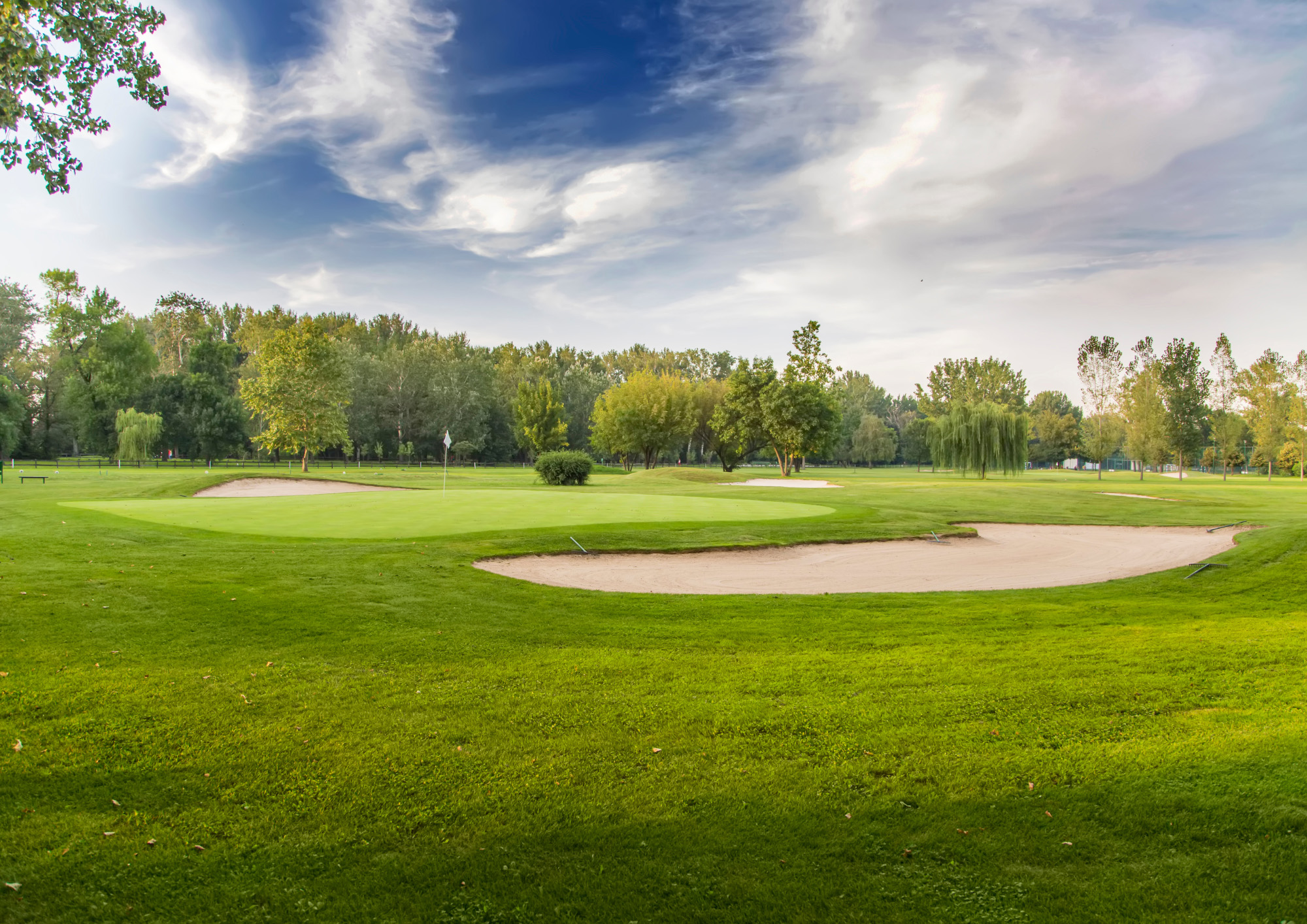 View of a golf course with green grass, sand bunkers, and a flag on the putting green, surrounded by trees under a partly cloudy sky.