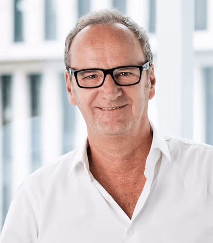 Mann mit Brille und weißem Hemd lächelt vor unscharfem Hintergrund mit Fenstern.