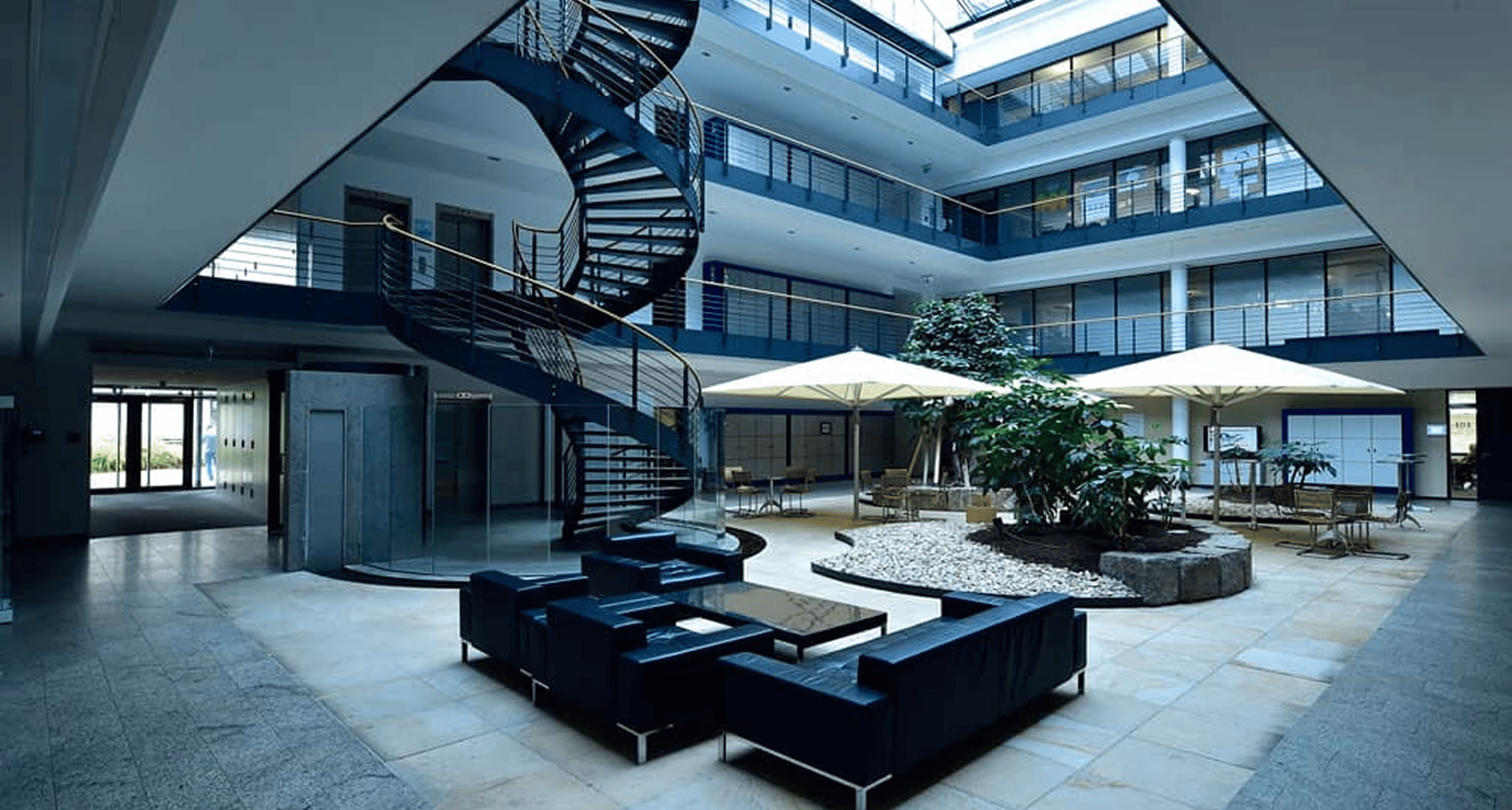 Moderne Bürolobby mit Wendeltreppe, schwarzer Ledercouch, großen Pflanzen und Glasdach für natürliche Beleuchtung.