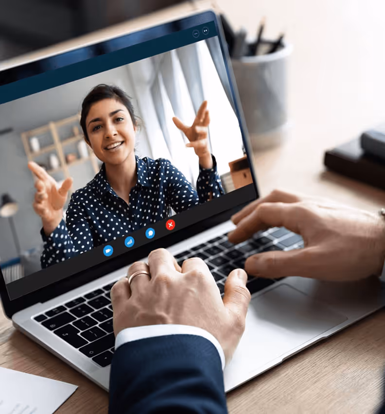 Frau im Videoanruf auf Laptop diskutiert, während jemand in Anzug am Gerät arbeitet. Büroatmosphäre mit Licht und Notizen.