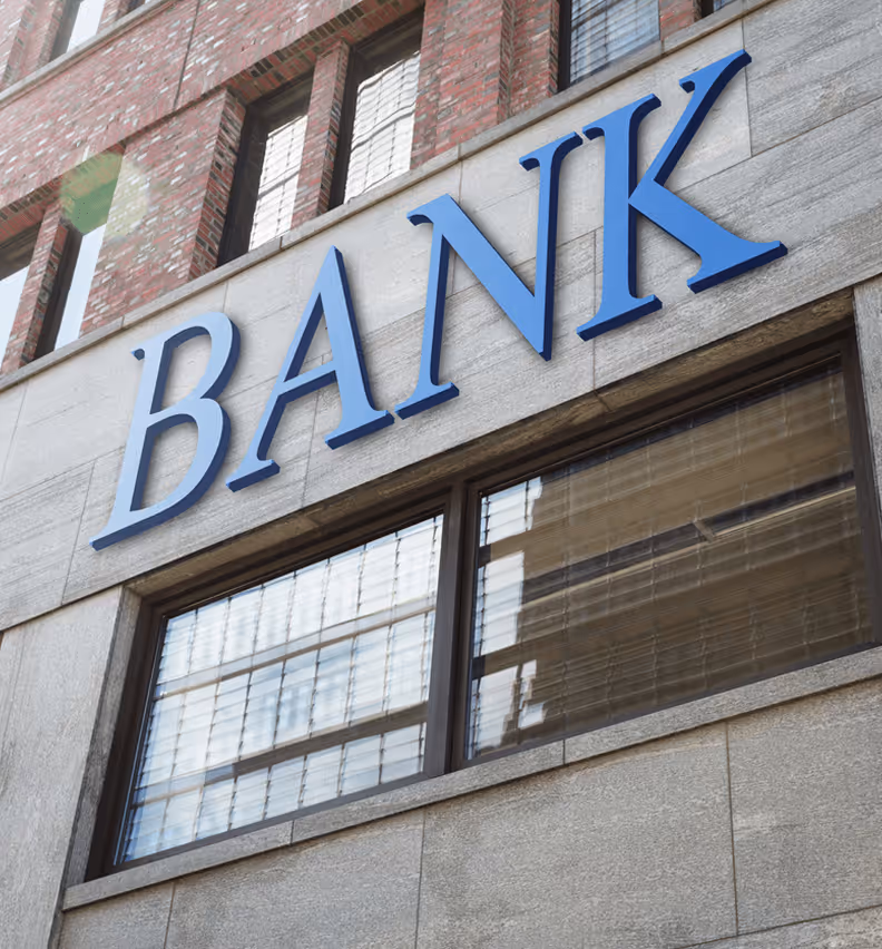 Moderne Bankfassade mit großem, blauem „BANK“-Schriftzug und rotem Backsteingebäude im Hintergrund.