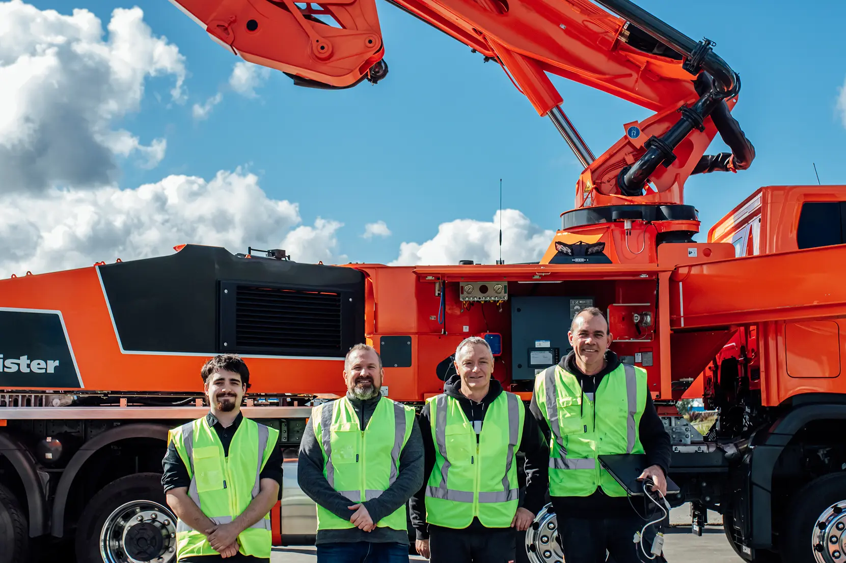 Four workers in neon safety vests stand in front of large orange industrial machinery under a blue sky.