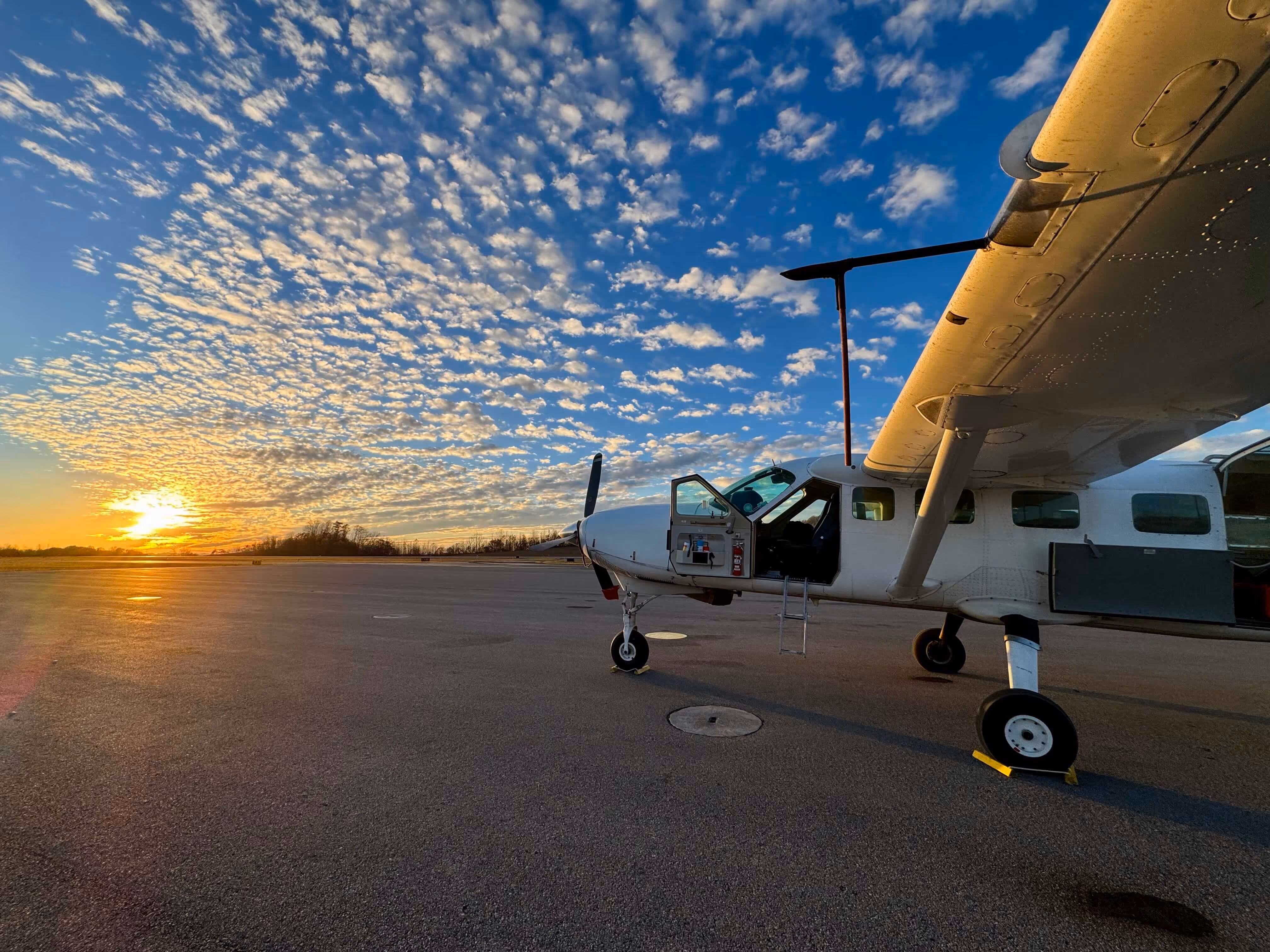 White Cessna Caravan airplane parked on runway at sunset with vibrant scattered clouds in blue sky.