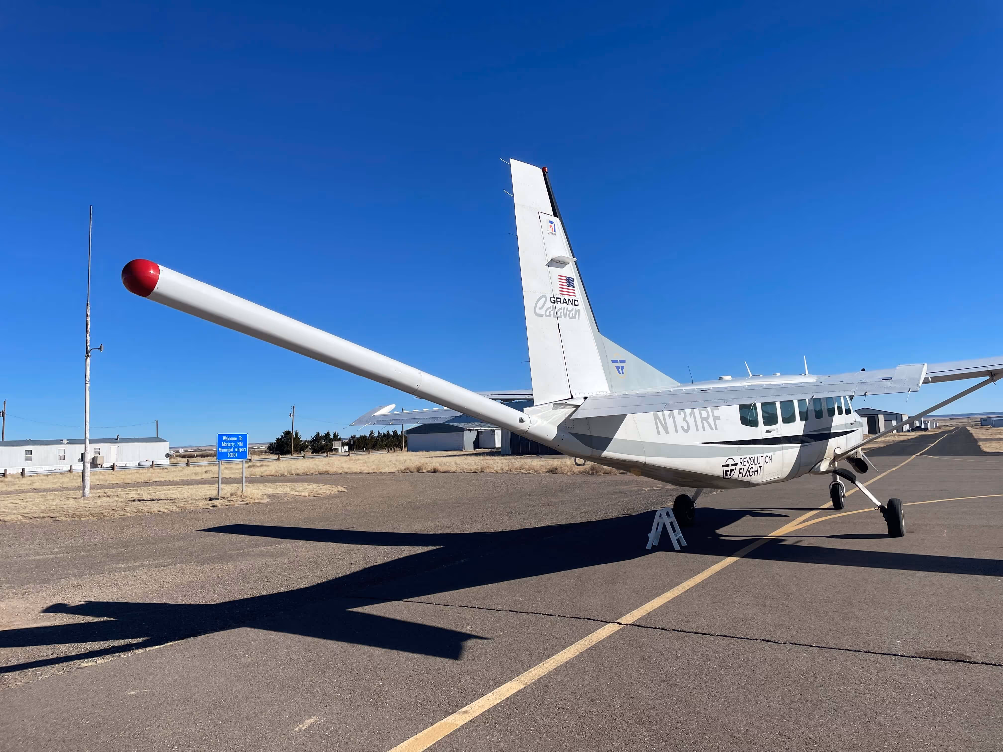 White Cessna Caravan parked on an asphalt runway under clear blue sky, with visible geophysical tail boom and hangars in the background.