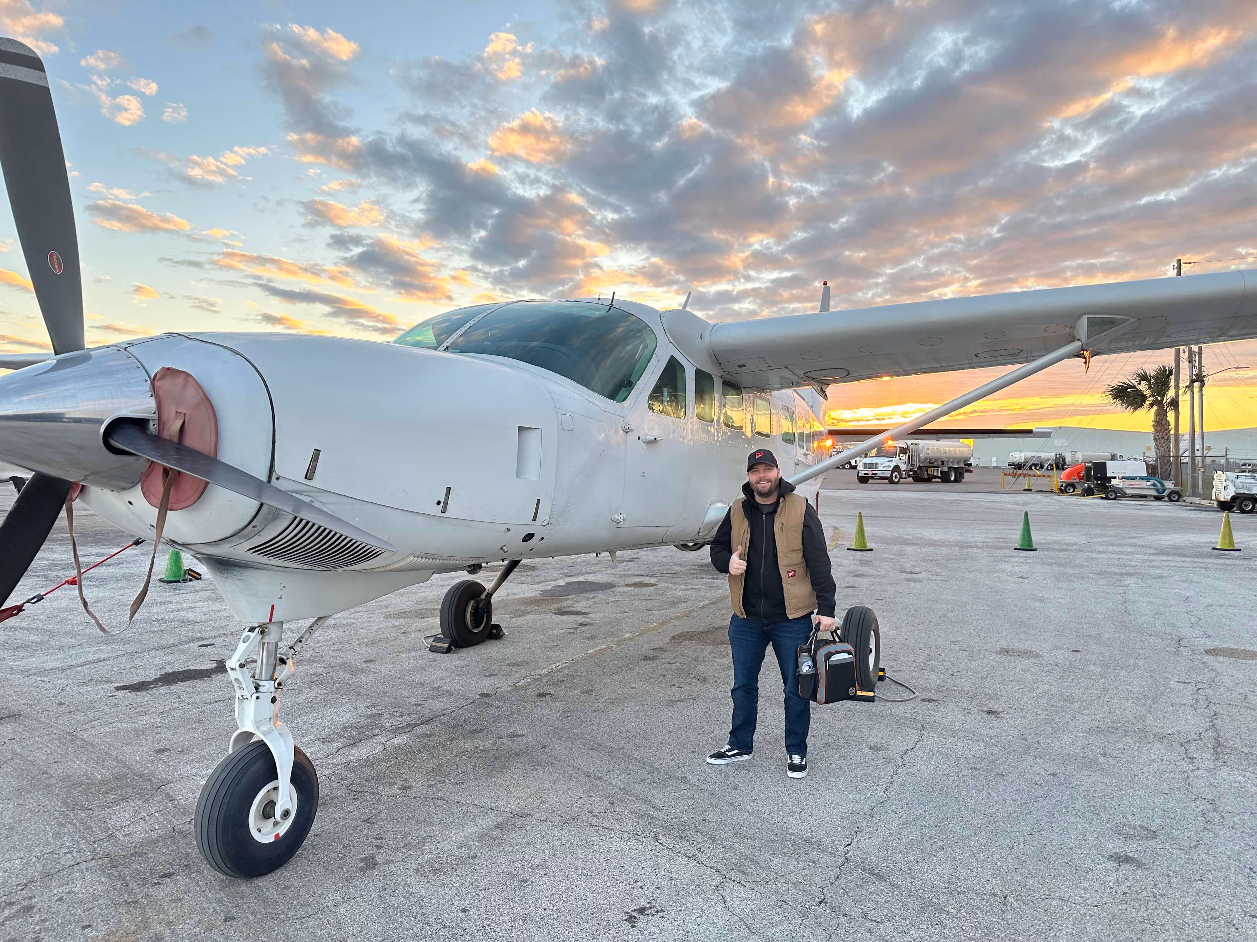 Man standing on airport tarmac next to a white Cessna Caravan during sunset.