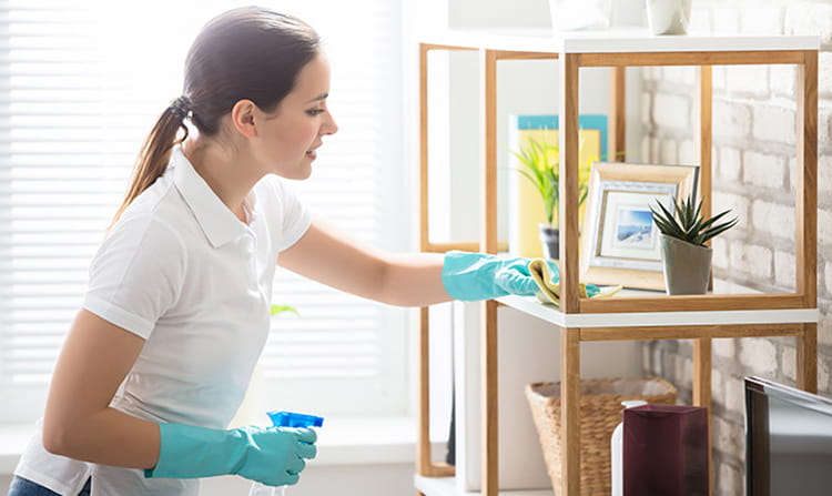 Woman wearing gloves cleaning a wooden shelf in a bright room with cleaning spray and cloth.