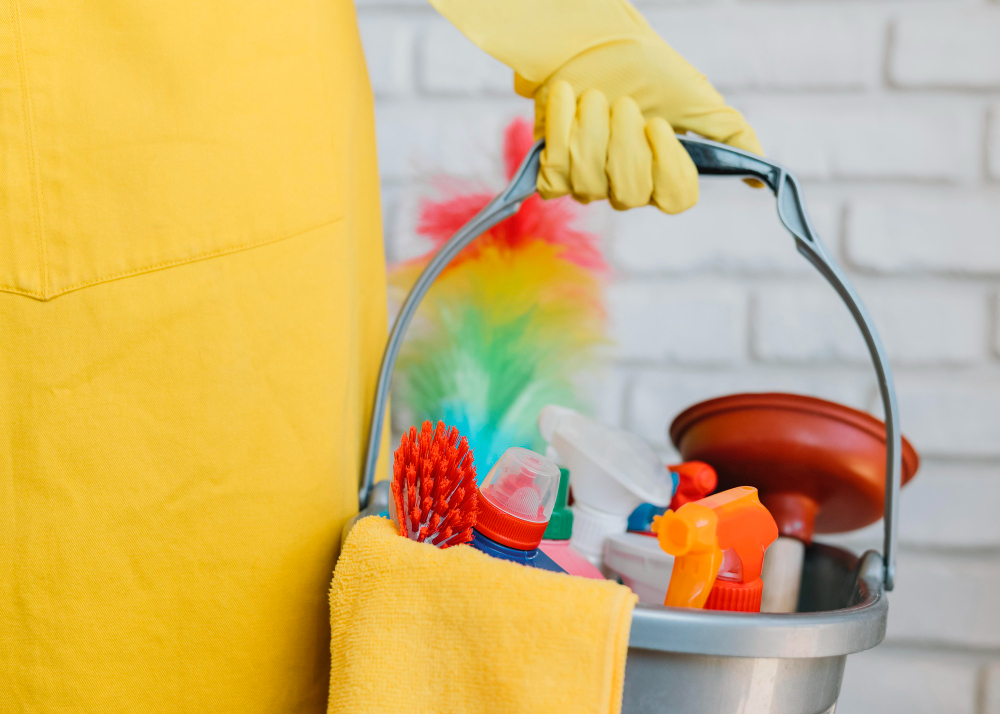 Person wearing yellow gloves and apron holding a bucket filled with cleaning supplies including brushes, spray bottles, a plunger, and a yellow cloth.