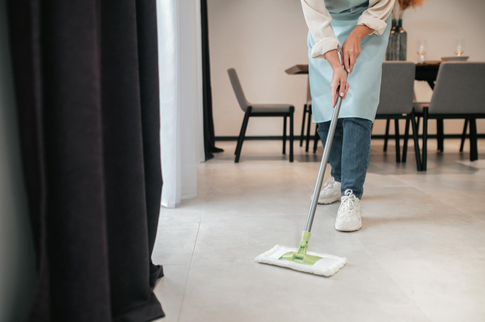 Person wearing an apron mopping a tiled floor in a living room with chairs and a dining table.
