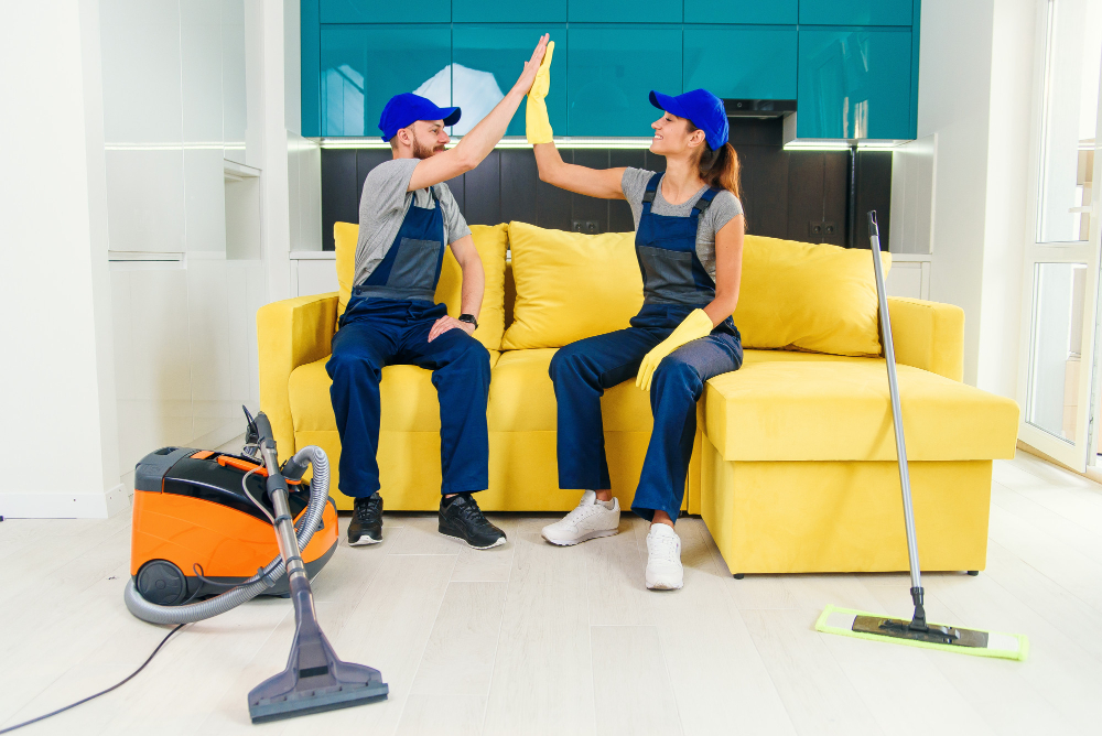 Two professional cleaners in uniform and gloves sitting on a yellow couch giving a high five with cleaning equipment on the floor.