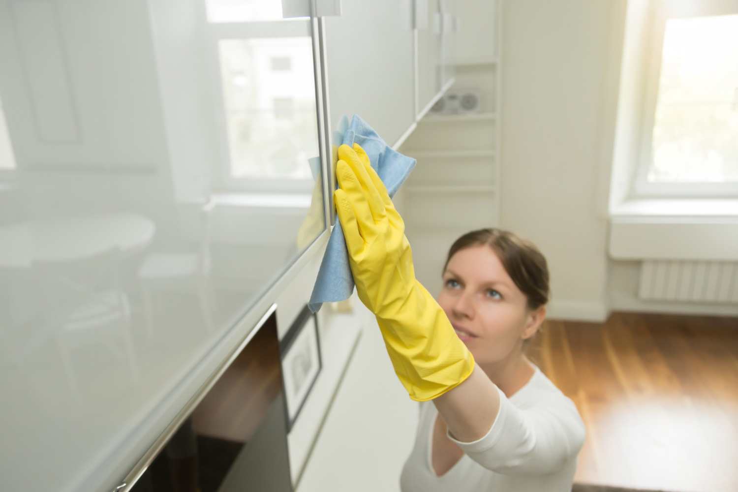 Woman wearing yellow rubber gloves wiping a glossy white cabinet with a blue cloth in a bright room.