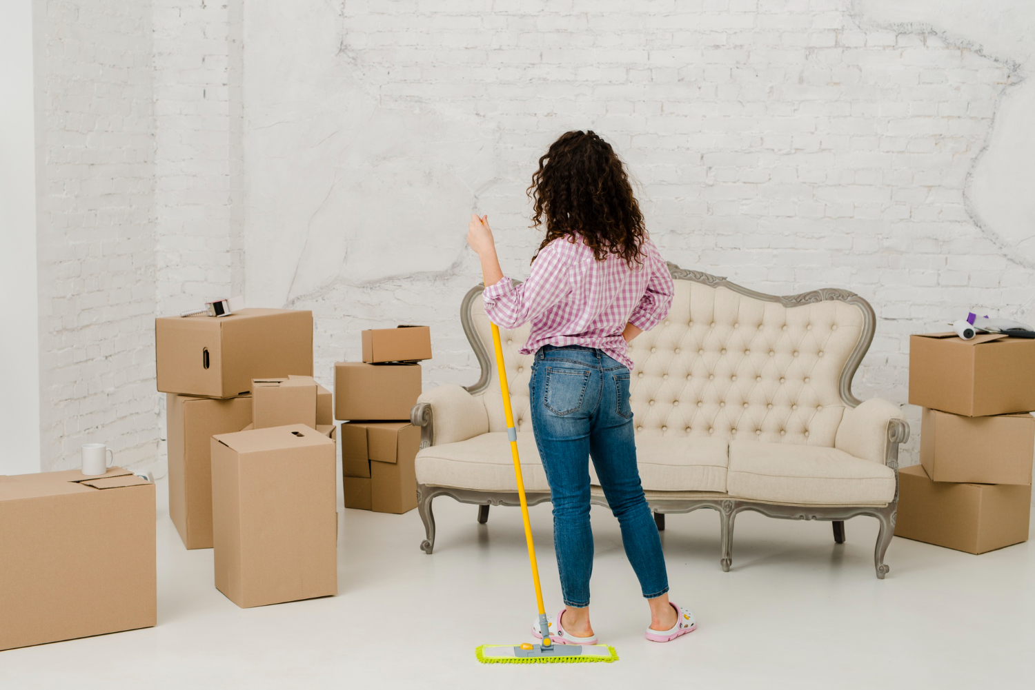 Woman with curly hair in jeans and a pink checkered shirt holding a mop, standing in a room with stacked cardboard boxes and a beige vintage sofa.