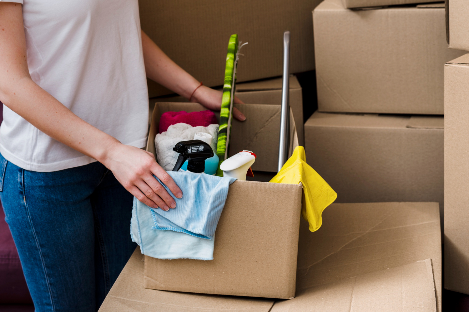 Person packing a cardboard box with cleaning supplies including spray bottles, cloths, and a mop amidst other packed boxes.