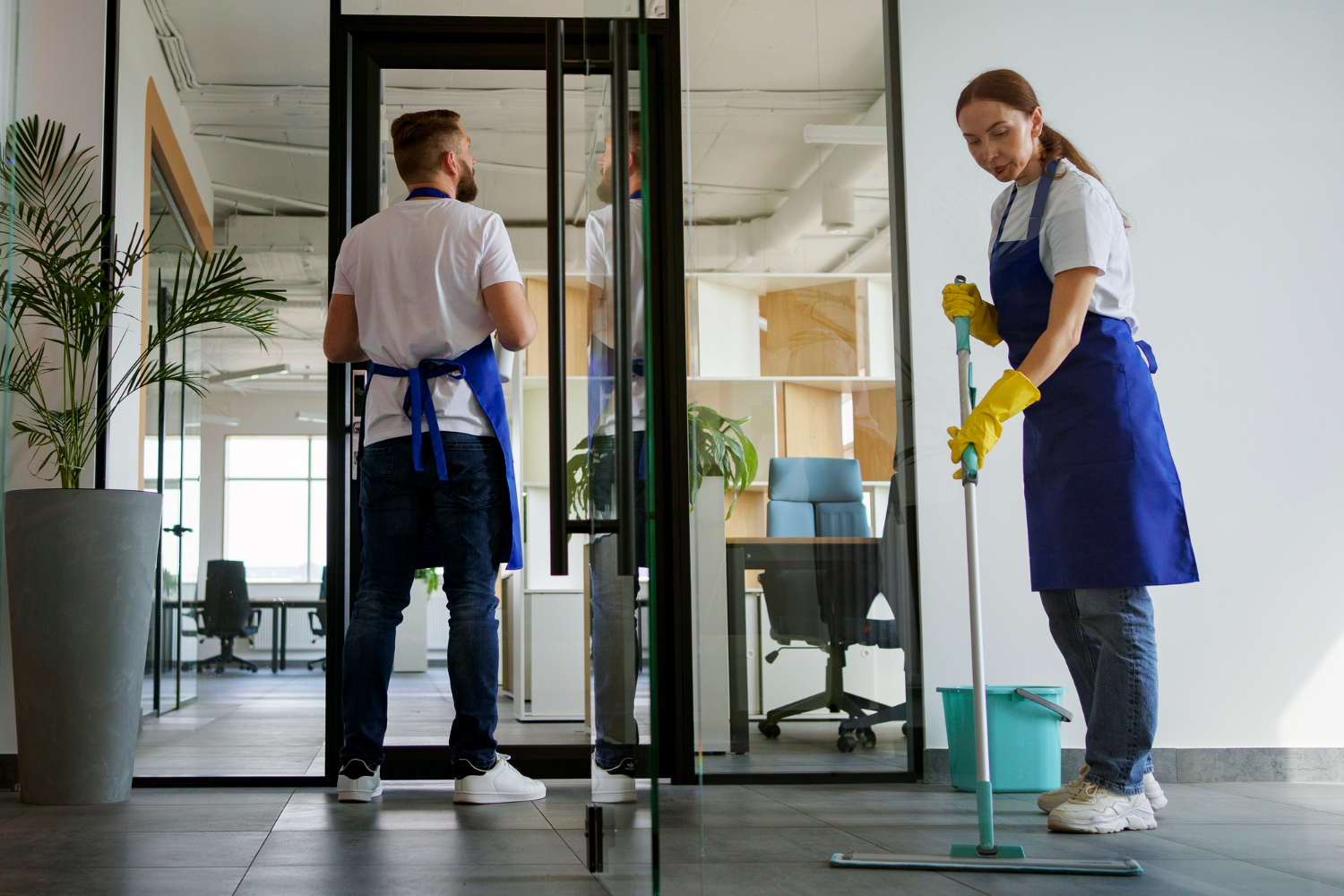 Two professional cleaners wearing blue aprons and yellow gloves cleaning an office; one mopping the floor and the other cleaning a glass door.