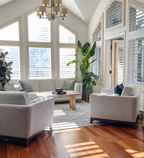 Bright living room with large windows, gray sofa and armchairs, wooden coffee table, green plants, and hardwood floor.