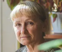 Portrait of an older woman with short blonde hair and a neutral expression in soft natural light.