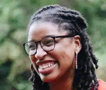 Smiling woman with dreadlocks, glasses, and hoop earrings outdoors with blurred green background.