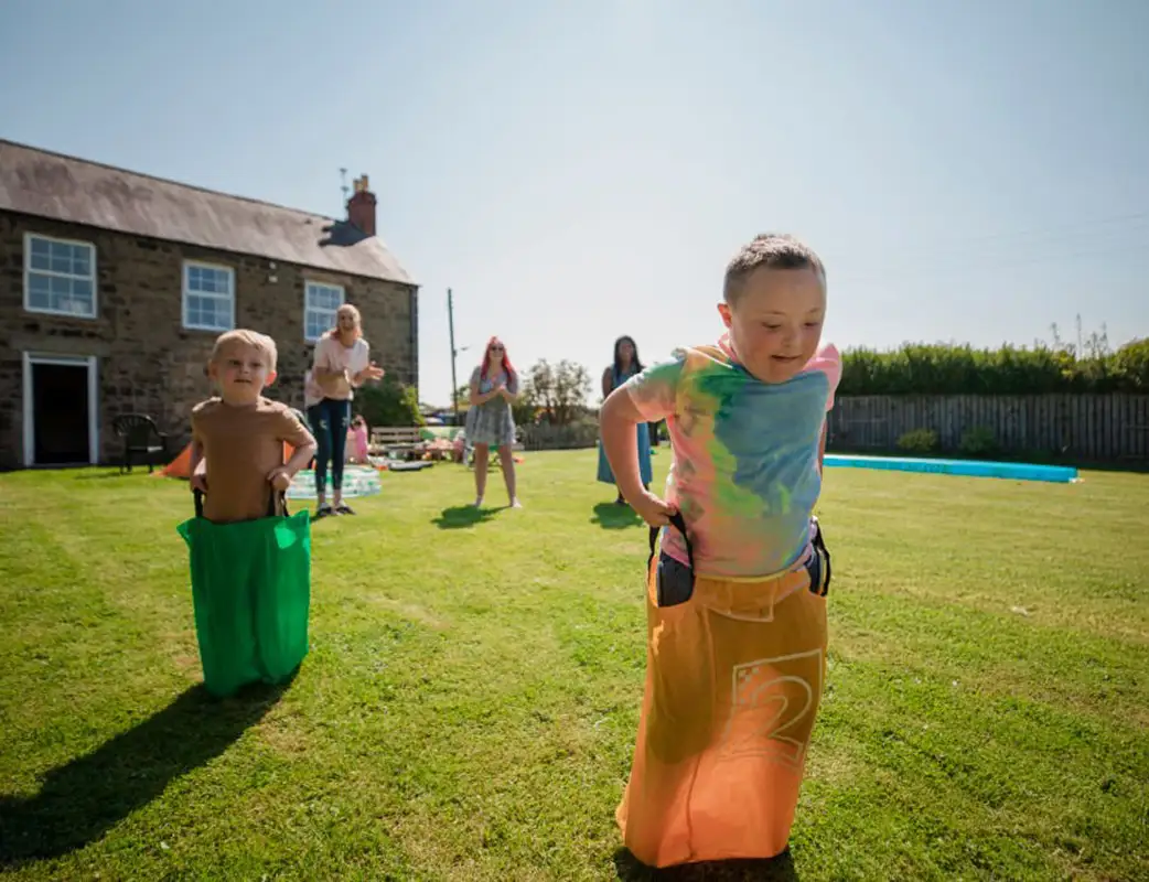 Two children participating in a sack race on a grassy lawn with a house and adults in the background.