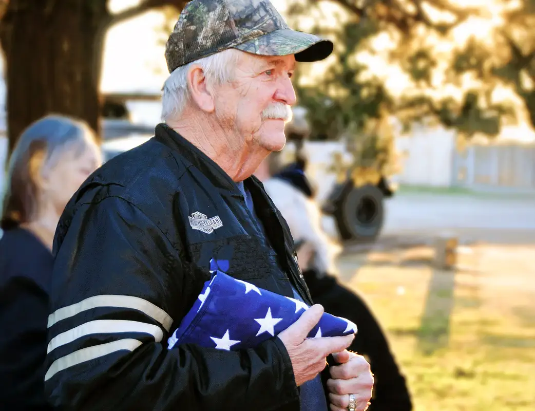 Older man wearing a camouflage cap and black jacket holding a folded American flag outdoors.