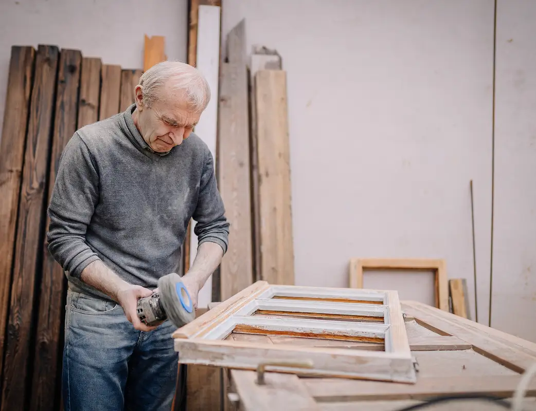 Older man using a power sander on a wooden window frame in a workshop.