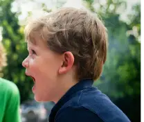 Young boy with light brown hair smiling and looking to the side outdoors with blurred green foliage background.
