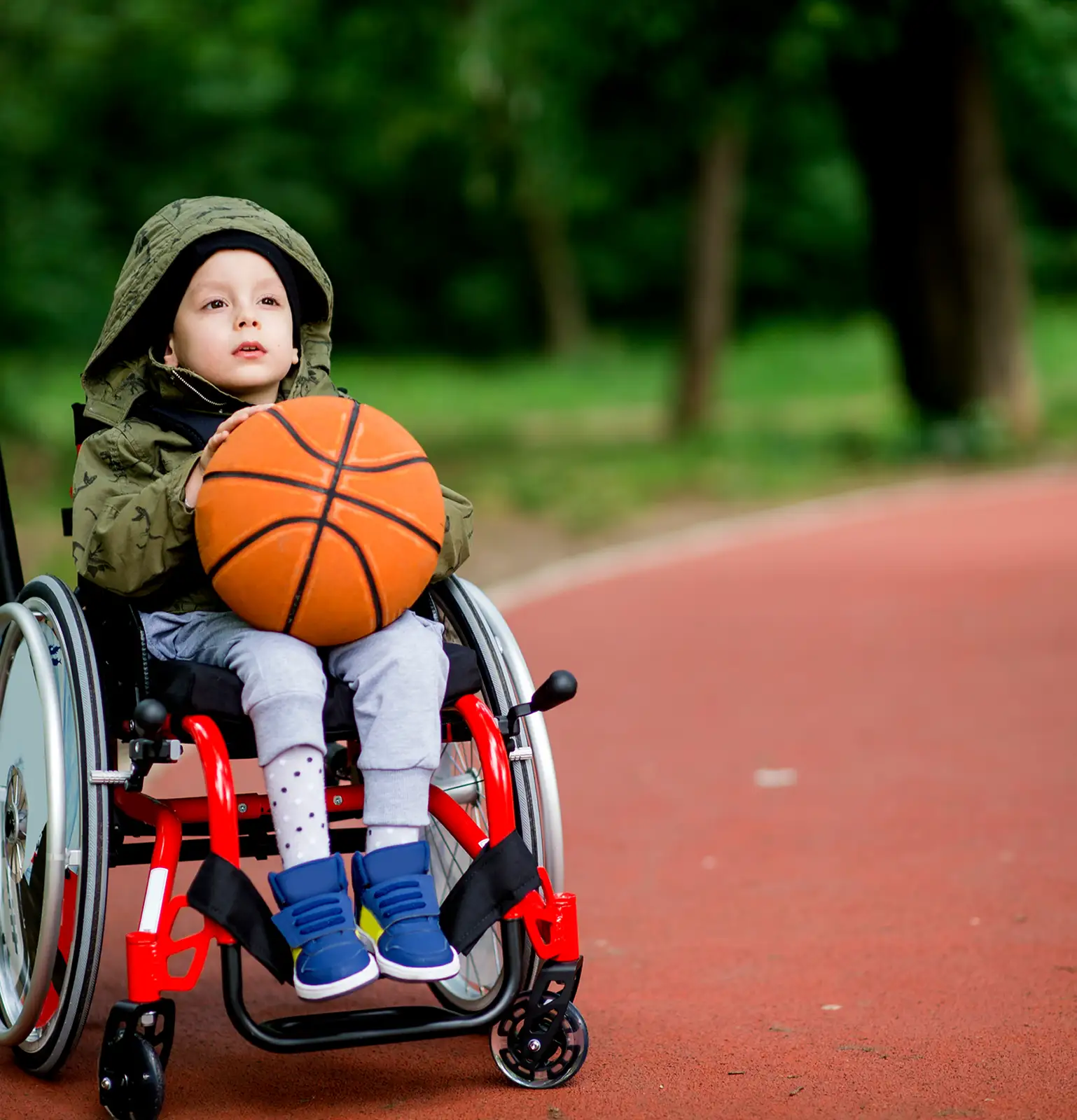 Child in a wheelchair holding a basketball on an outdoor sports court.