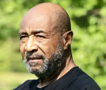 Portrait of an older bald man with a beard in a black shirt outdoors with greenery in the background.