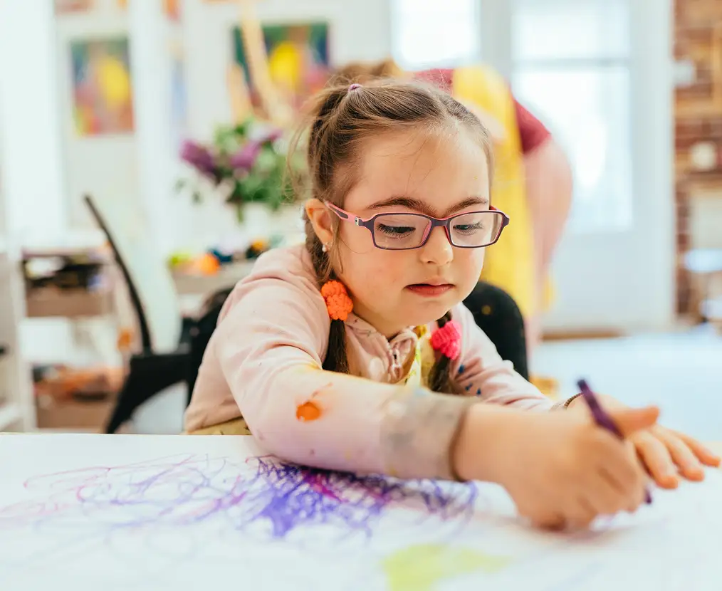 Young girl with glasses and braided hair coloring with a purple crayon on paper at a table.