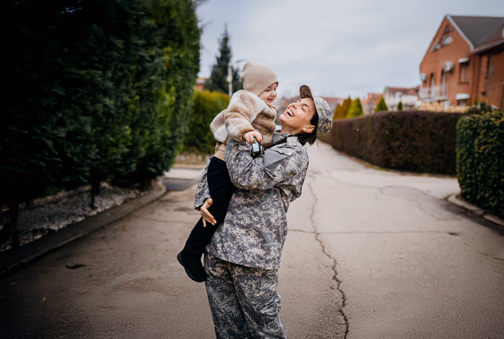 Smiling man wearing a green apron holding a tall leafy houseplant near a window.