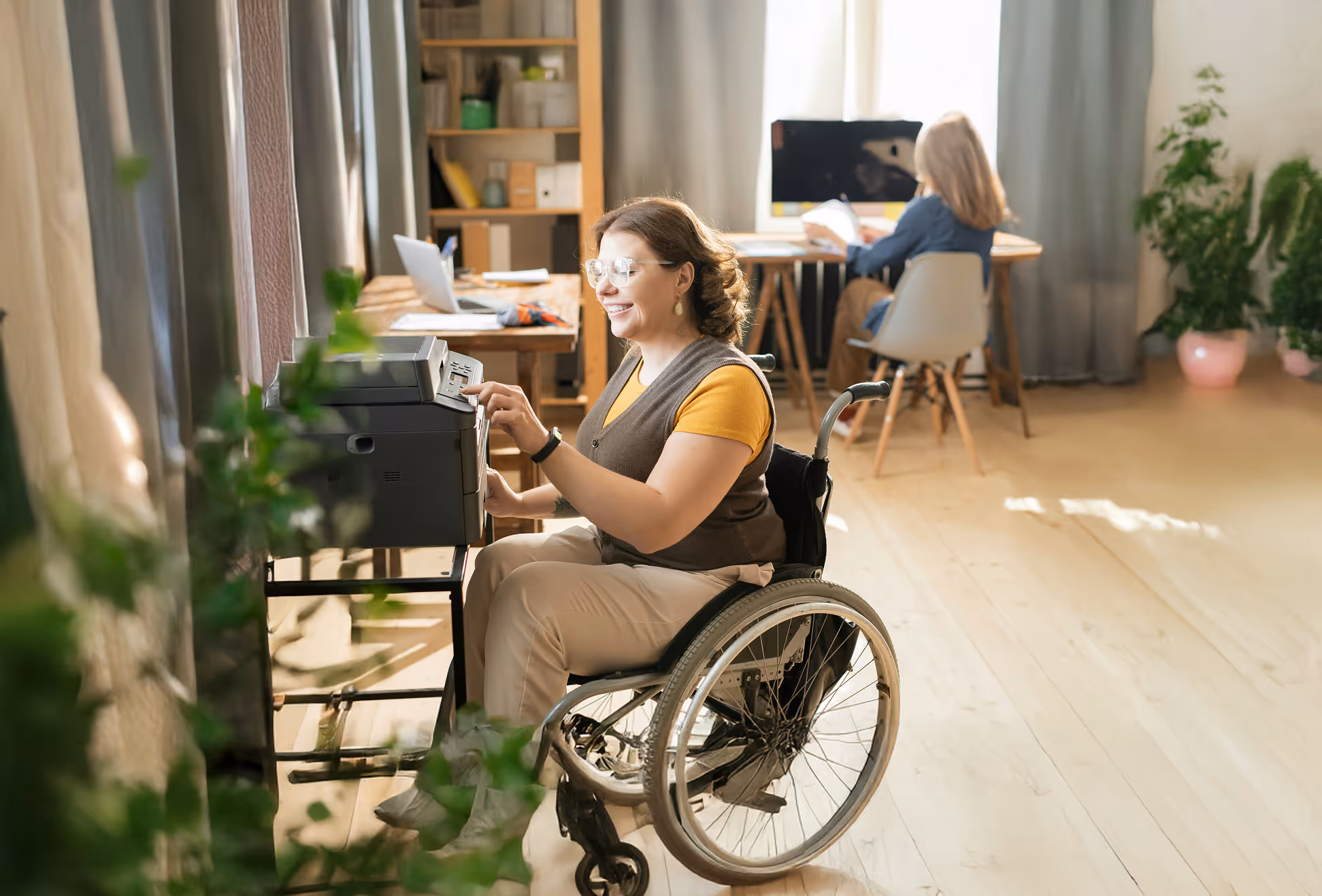 Smiling man wearing a green apron holding a tall leafy houseplant near a window.
