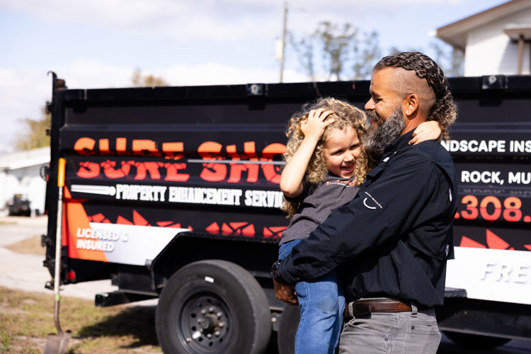 Christopher holding his daughter in front of a Sure Shot Property Enhancement Services trailer