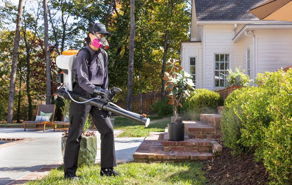 Person wearing mask uses a backpack sprayer to spray plants near a white house and garden.