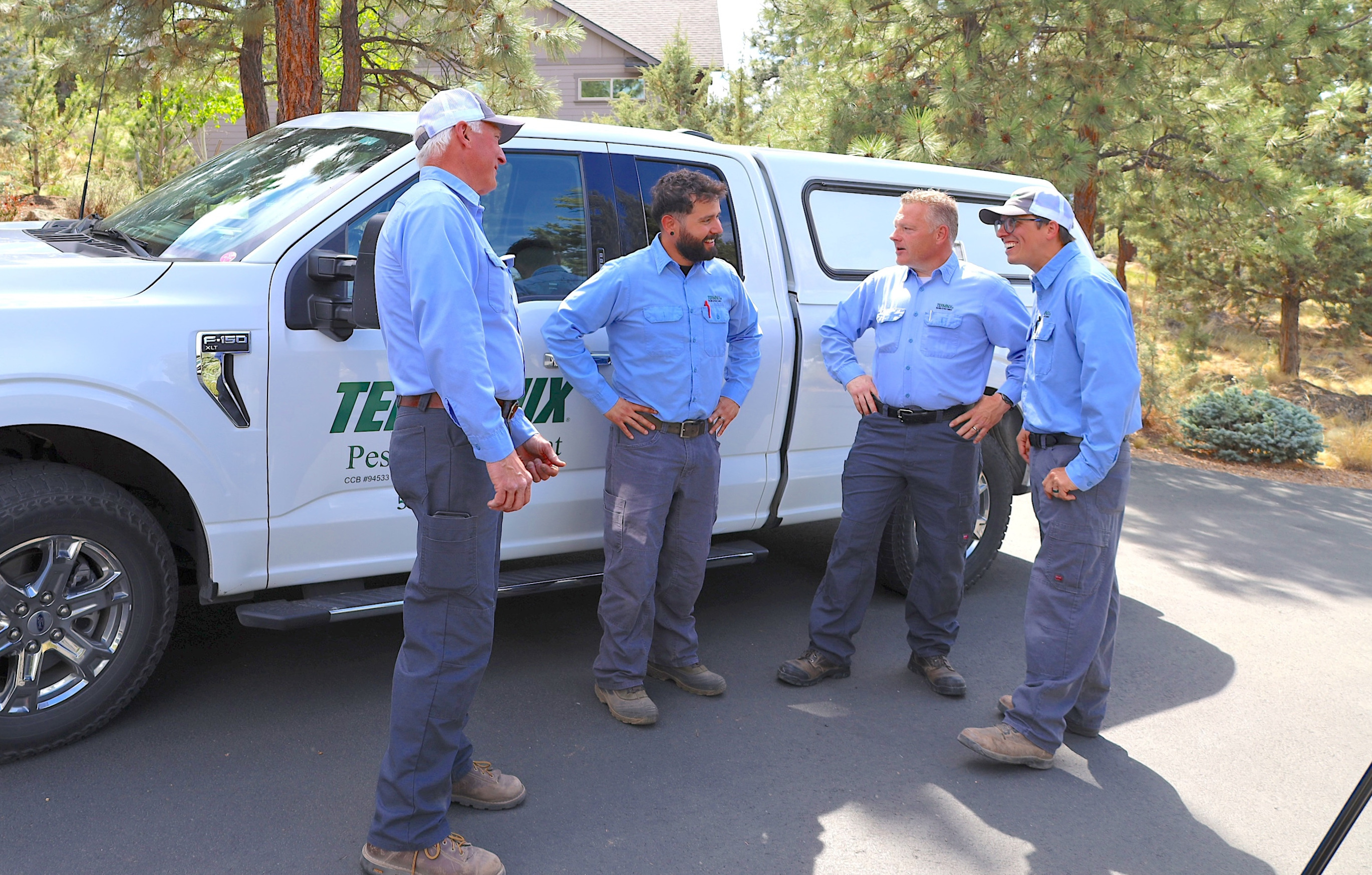 Four technicians in blue uniforms standing and talking beside a white service truck in a wooded area.