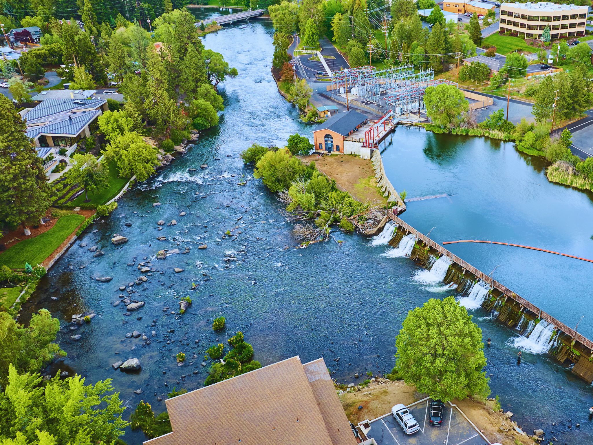 Aerial view of a river flowing past a dam, surrounded by trees, buildings, and a parking lot.