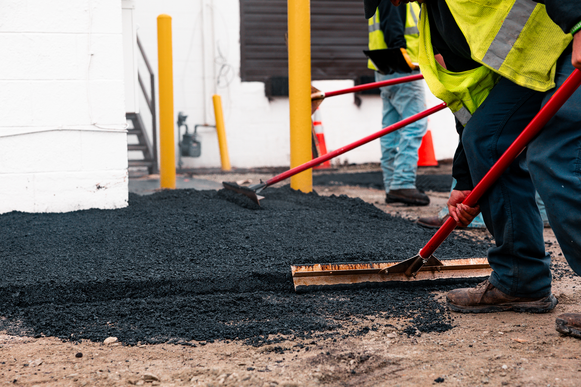 Construction workers in safety vests spreading and leveling freshly laid asphalt on a ground surface near a building.