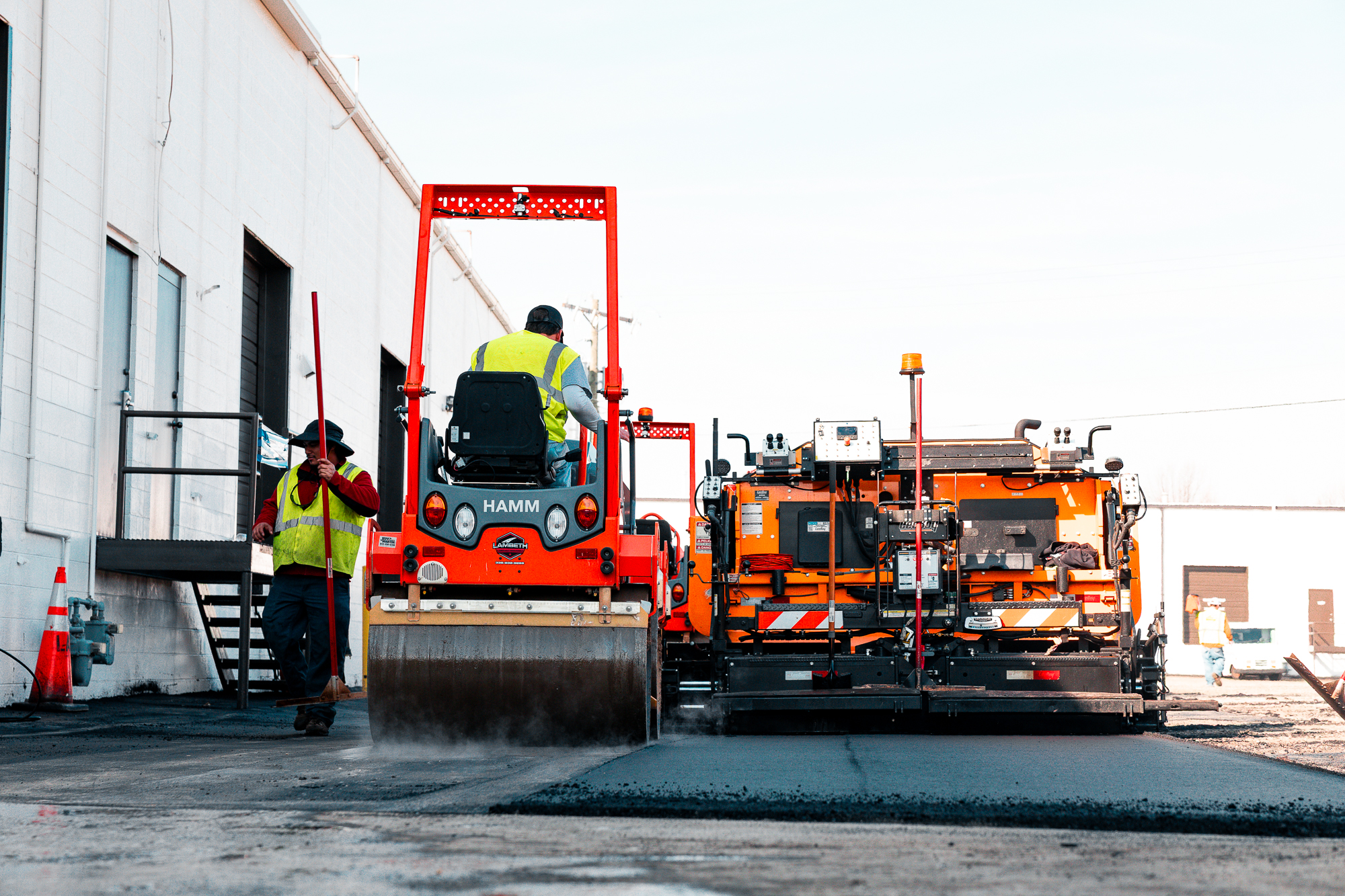 Two construction workers in safety vests operating heavy machinery to pave a road beside a white industrial building.
