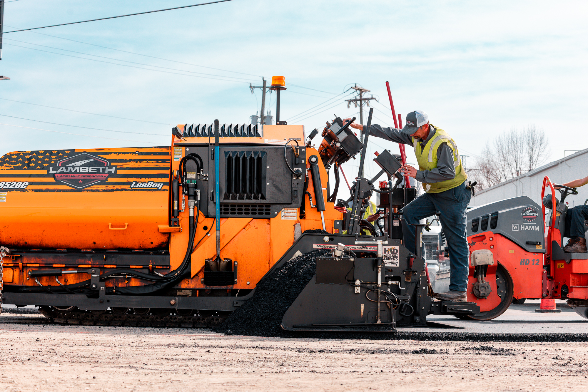 Construction worker operating an orange asphalt paving machine labeled Lambeth Asphalt Services on a road site.