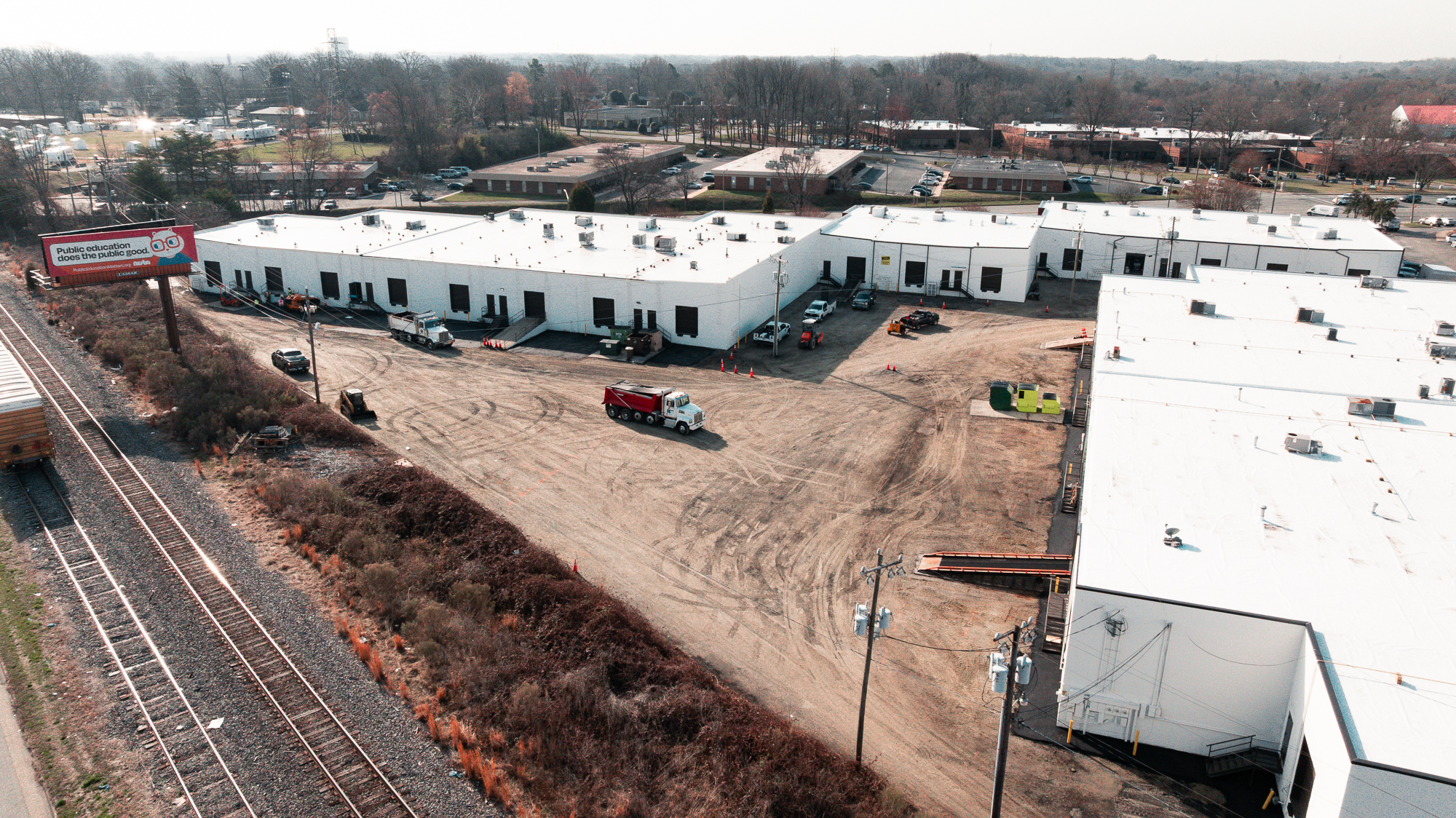 Aerial view of industrial buildings with white roofs, dirt parking lot with trucks and construction equipment, next to railroad tracks and a billboard.