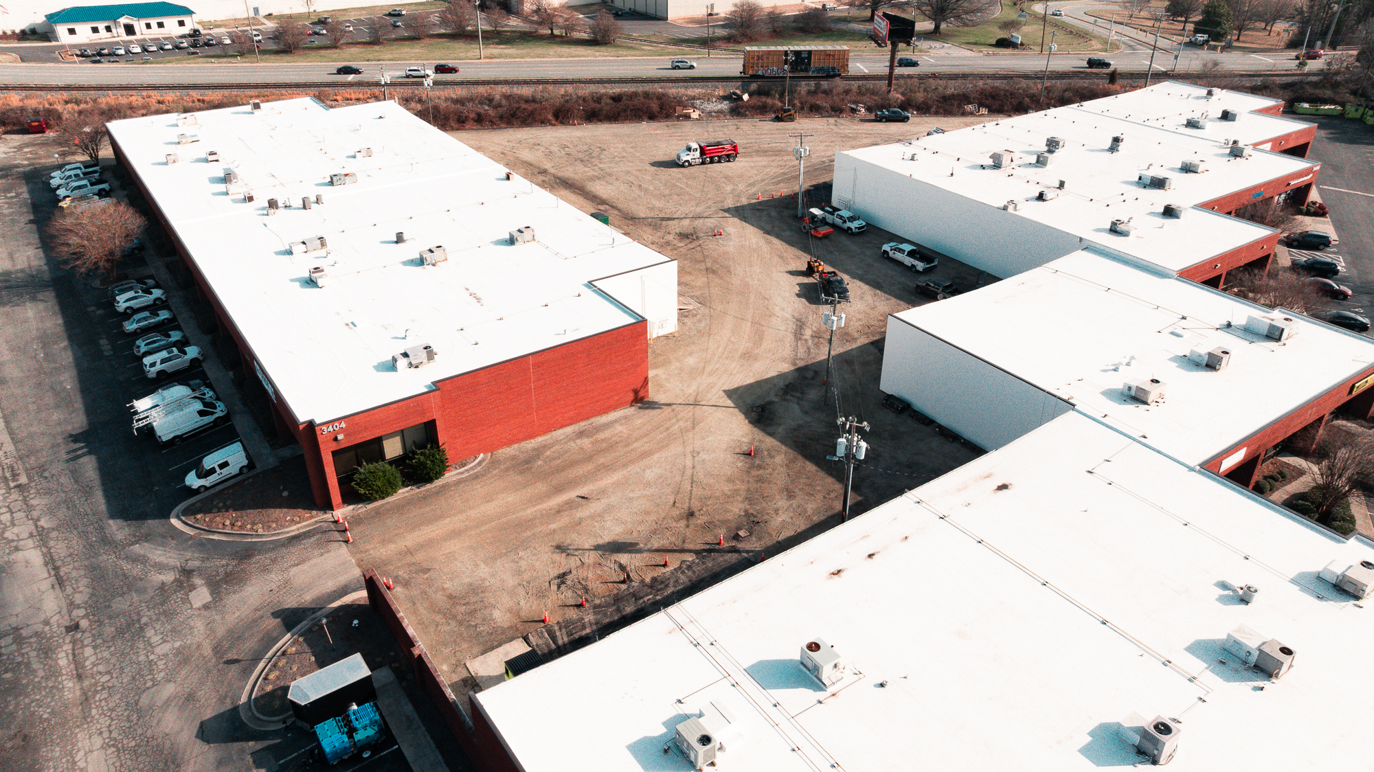 Aerial view of commercial warehouse buildings with white roofs, surrounded by trucks and vehicles in a parking area.