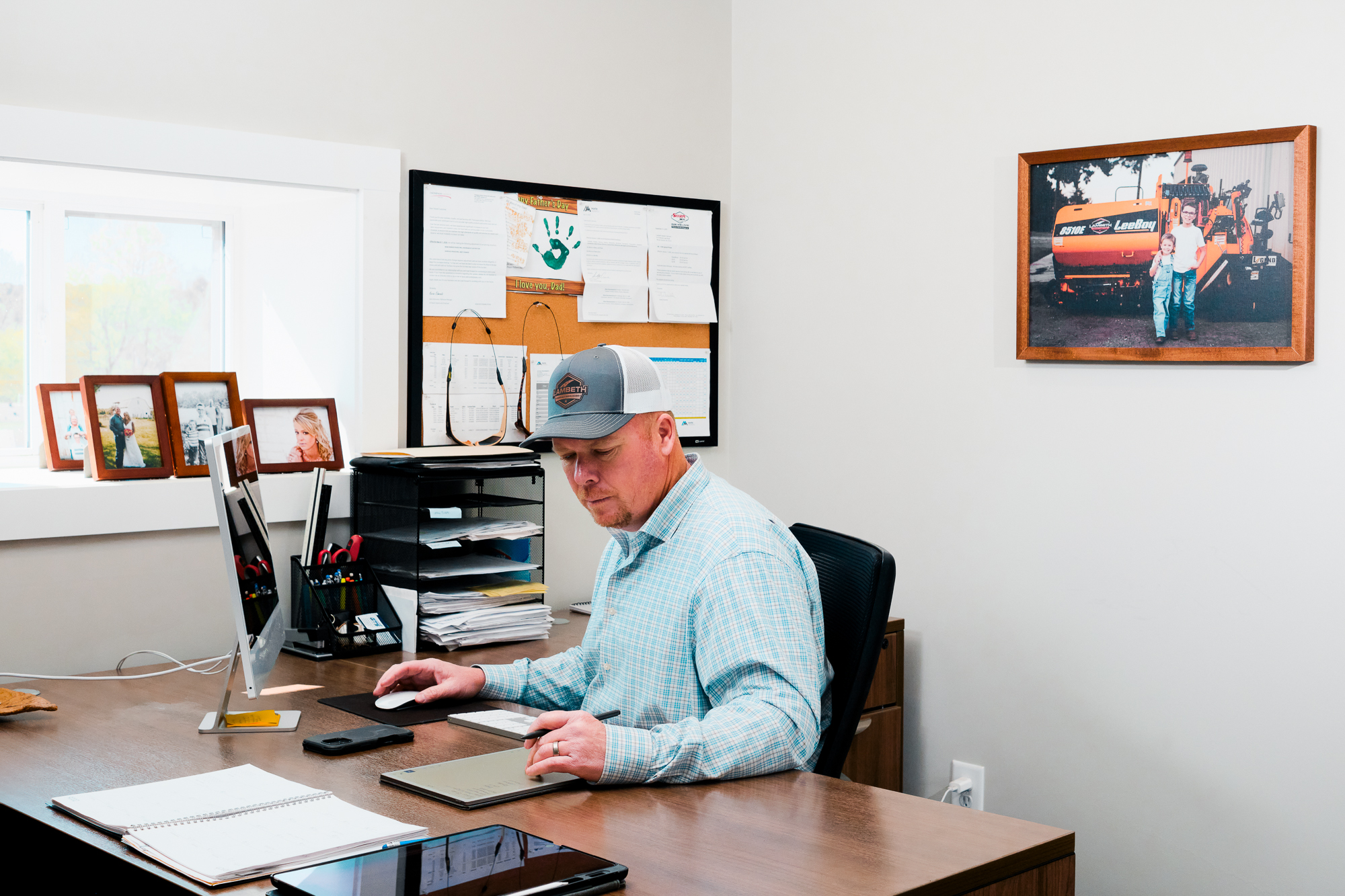 Man in a plaid shirt and cap using a digital pen on a tablet at a desk with a desktop computer and family photos in the background.